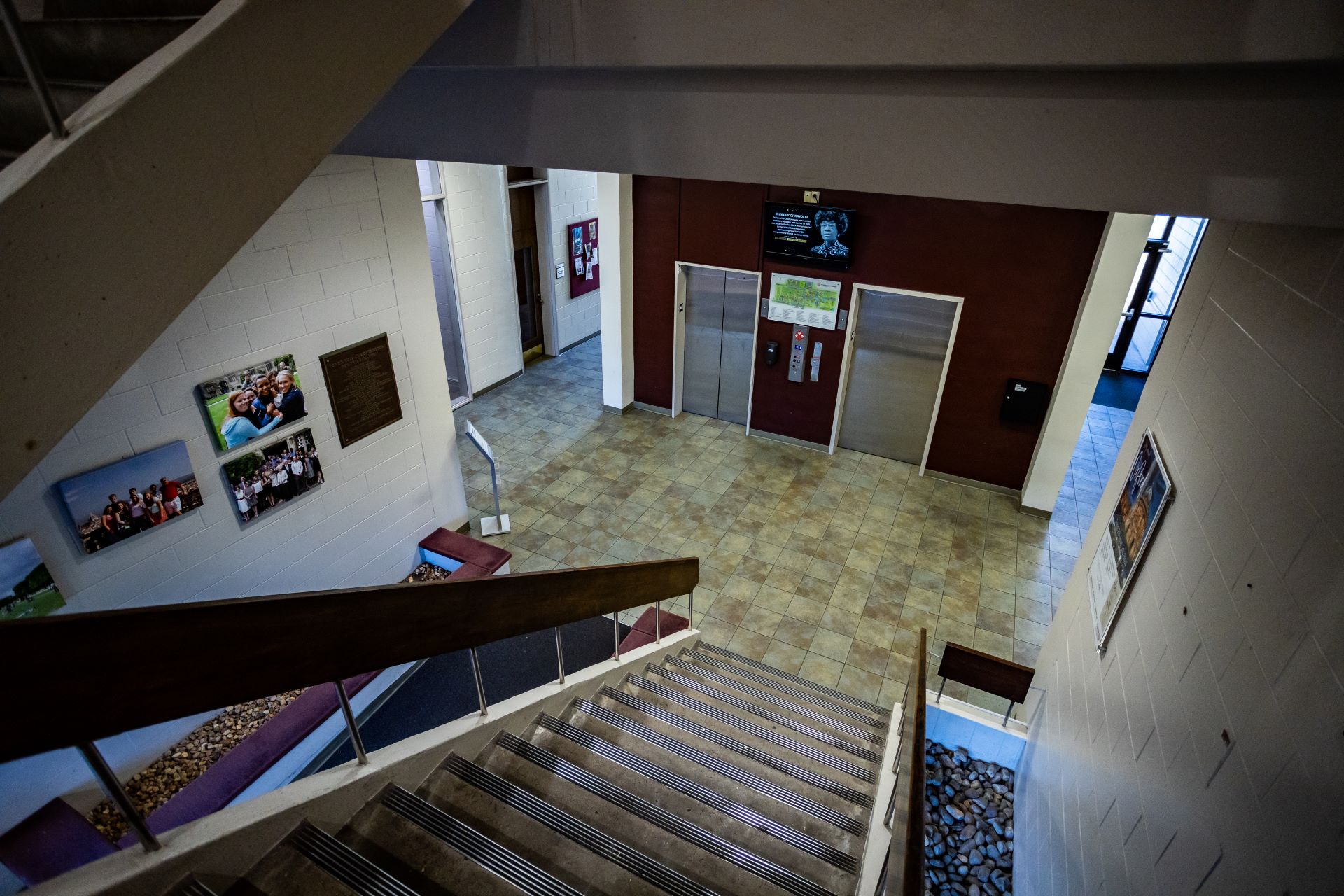 Overhead view of a building's lobby showing a staircase descending towards two elevators, with walls decorated with framed photographs